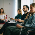 Teilnehmer sitzen auf Stuehlen nebeneinander und verfolgen das Coaching