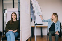 Eine Teilnehmerin macht Notizen auf einer Flipchart, eine Frau sitzt durch ein Whiteboard getrennt daneben