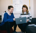 Zwei lächelnde Frauen sitzen vor einem Laptop und sehen auf den Bildschirm 